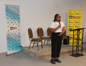 Woman in a white top and black pants speaks into a microphone on a small stage, with JA Bank and Money banners nearby.
