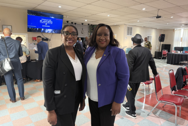 Two smiling women in black and purple blazers pose for a photo at an indoor event, with others in the background and a screen showing 'Stanley Cup Playoffs'.