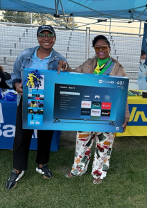 Two women under a blue canopy hold a boxed 40-inch Philips Roku TV prize at an outdoor event.